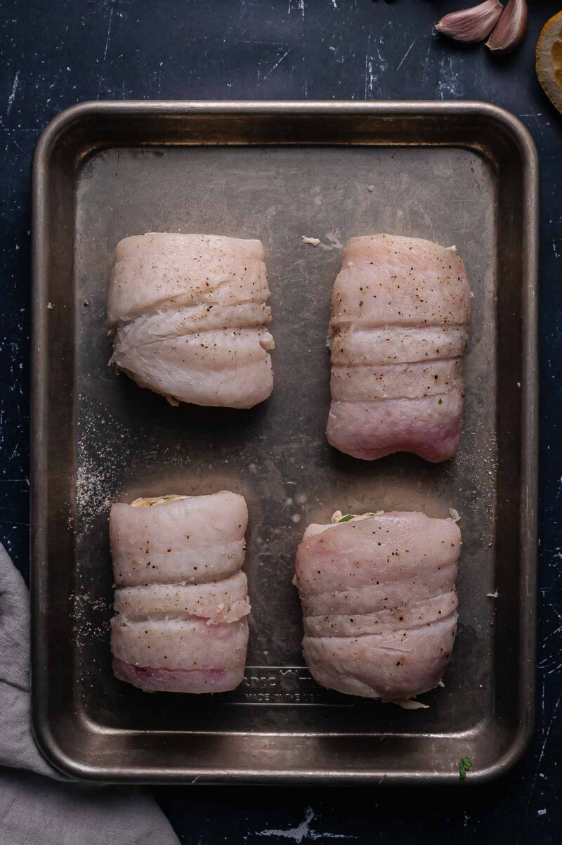 A metal baking tray with four uncooked, seasoned fish fillets rolled and spaced evenly apart. Garlic cloves and a lemon slice are partially visible in the corners of the image.