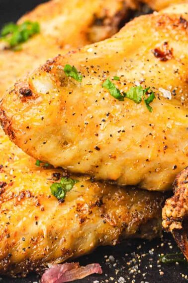 A close-up of seasoned and baked chicken wings garnished with chopped herbs, served on a dark plate with lemon wedges.