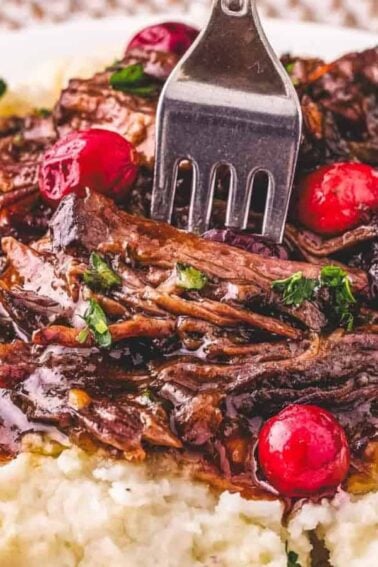 A plate of mashed potatoes topped with shredded pot roast, brown gravy, and garnished with red cranberries and chopped herbs.