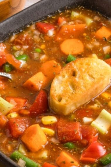 A bowl of vegetable soup with diced carrots, corn, green beans, potatoes, and tomatoes, topped with a slice of toasted bread and a spoon on the side.