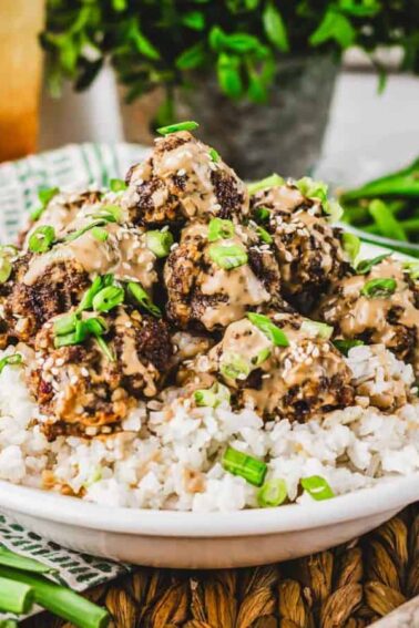 A bowl of white rice topped with meatballs covered in creamy sauce and chopped green onions, with green beans and fresh herbs in the background.