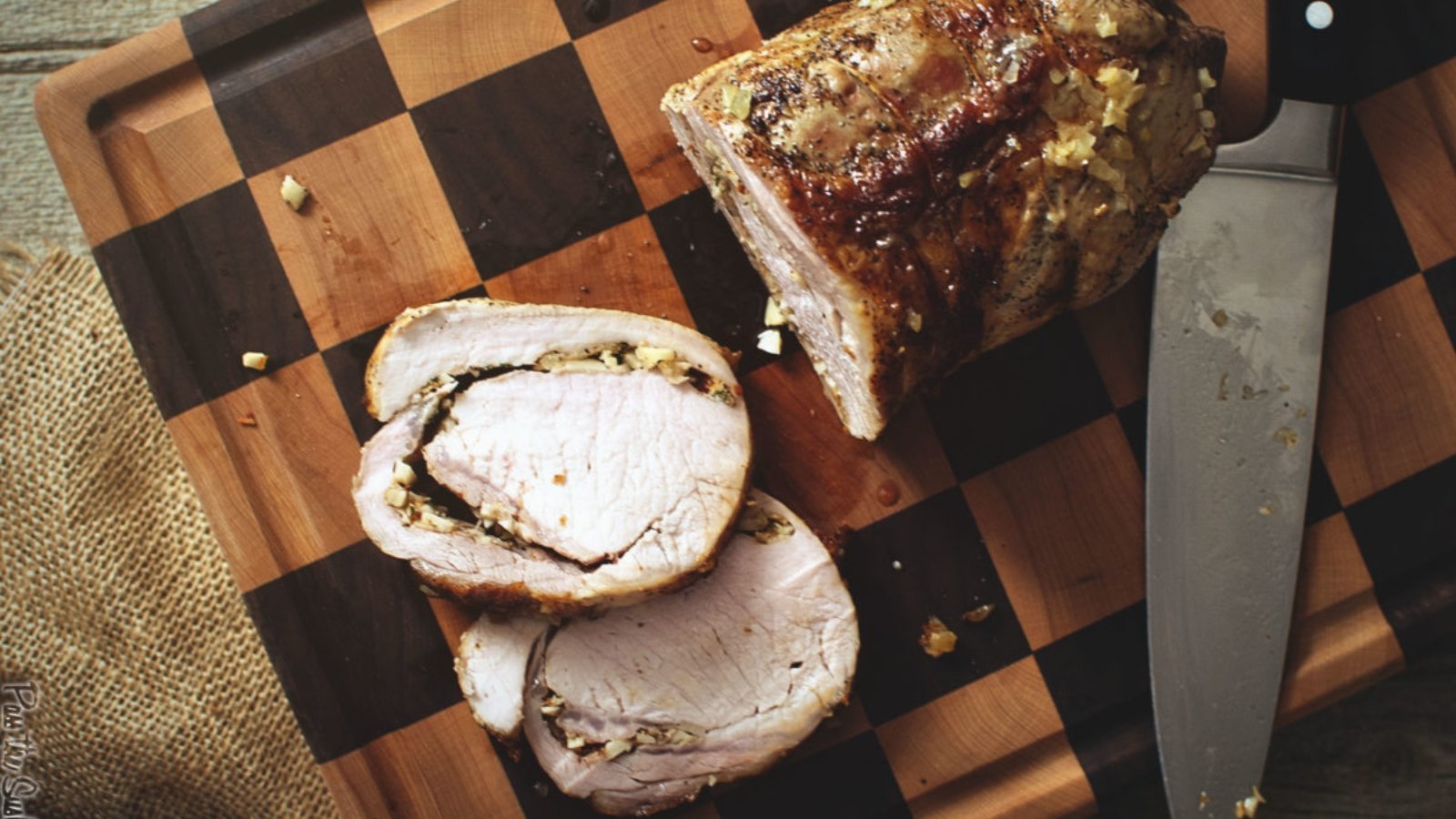 Roasted pork loin, partially sliced, on a checkered wooden cutting board with a knife beside it.
