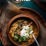 Overhead view of a bowl of green chicken chili topped with sour cream, jalapeños, and herbs, with a spoon on the side and text about making green chicken chili.