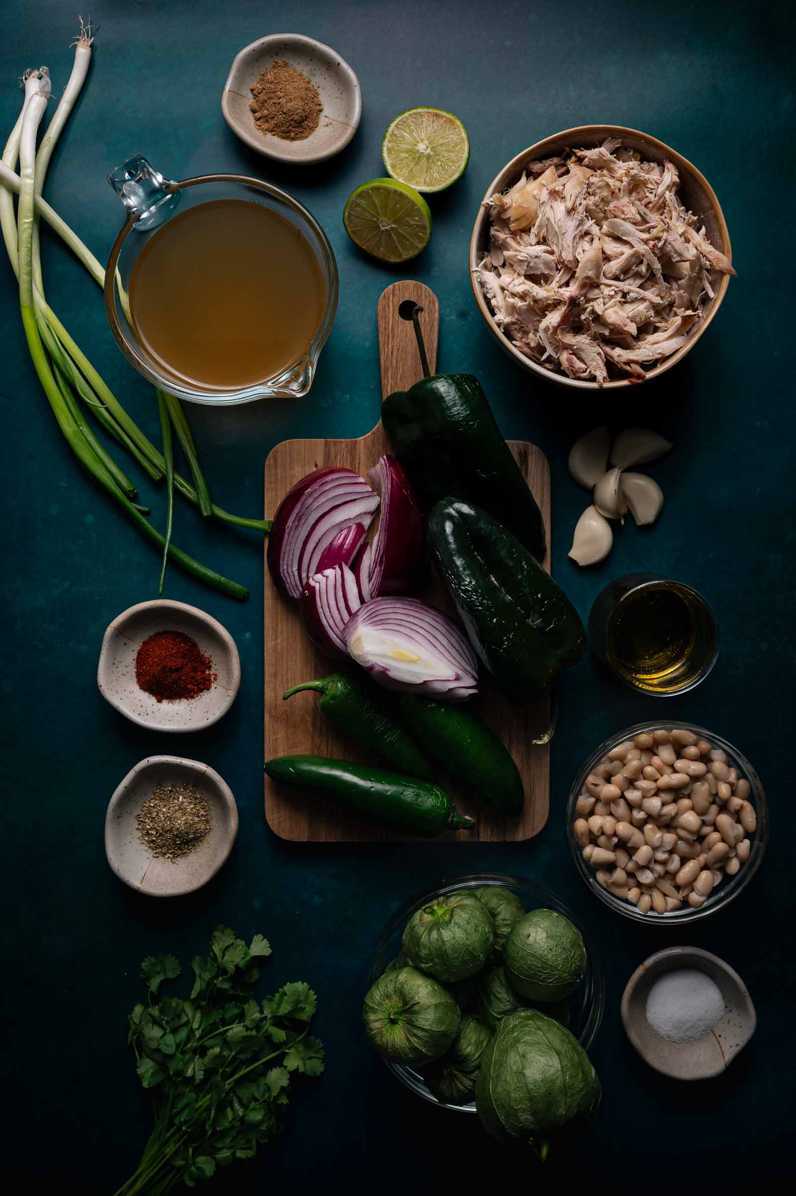 Flat lay of chicken, white beans, tomatillos, green onions, broth, red onion, garlic, cilantro, green chilies, dried spices, lime, and oil arranged on a dark surface.