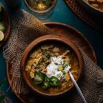 A wooden bowl of green chicken chili garnished with sliced jalapeños, sour cream, herbs, and tortilla chips sits on a wooden plate with a spoon, next to a drink and another dish.