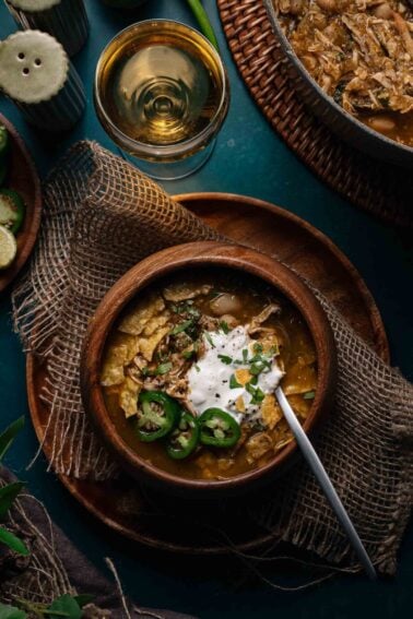 A wooden bowl of green chicken chili garnished with sliced jalapeños, sour cream, herbs, and tortilla chips sits on a wooden plate with a spoon, next to a drink and another dish.