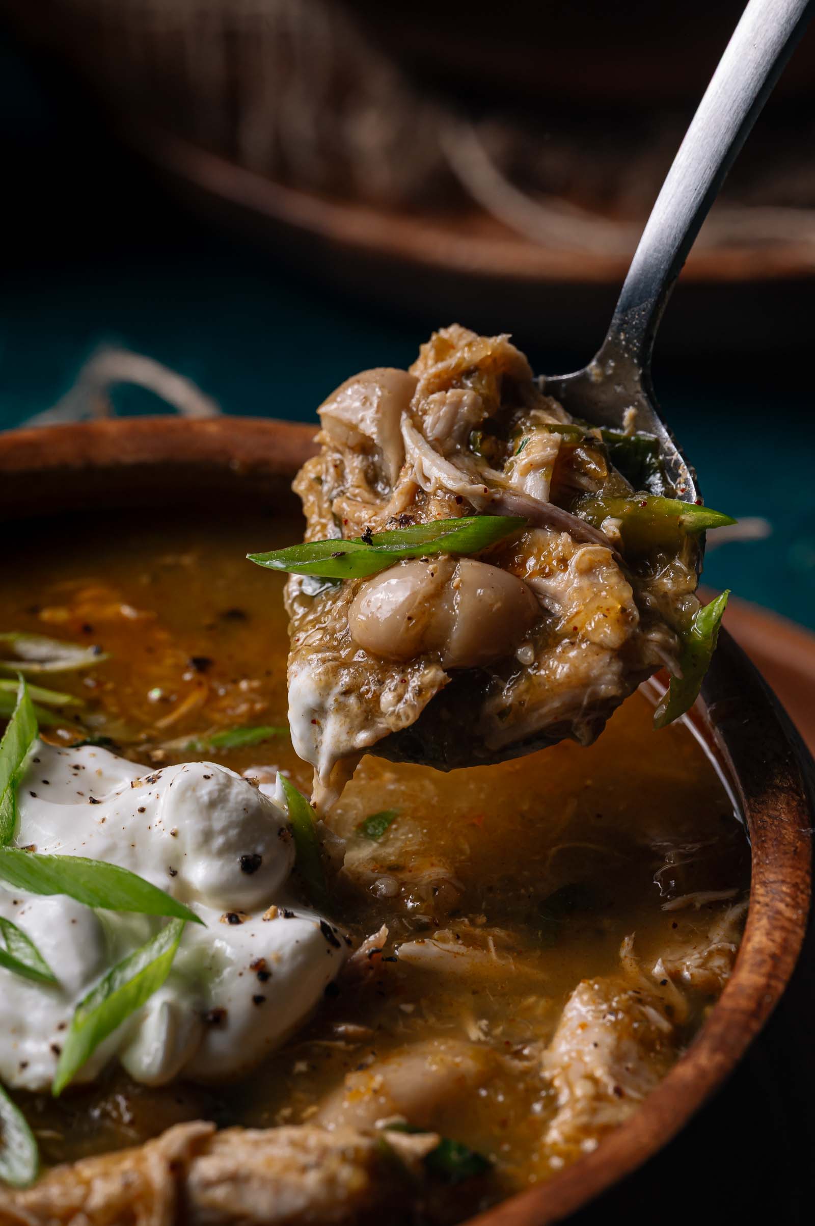 Close-up of a bowl of green chicken chili with shredded chicken, white beans, sliced green onions, and a dollop of sour cream; a spoon holds a portion above the bowl.