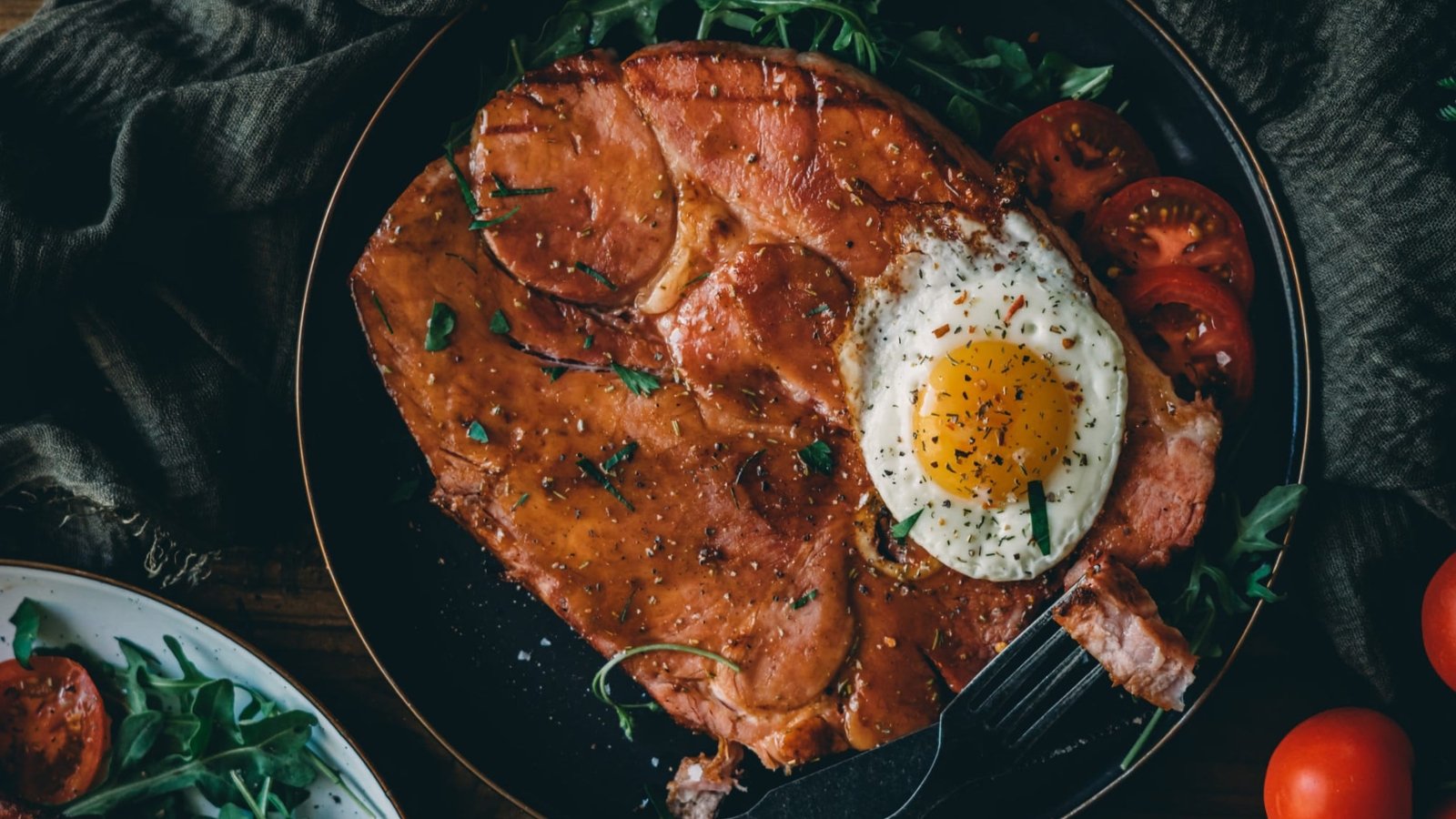 A plate with a large slice of grilled ham, a sunny-side-up fried egg, sliced tomatoes, and fresh greens.