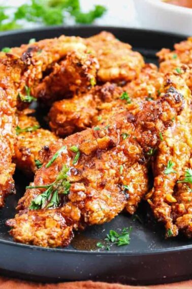 A black plate filled with crispy, breaded chicken tenders garnished with chopped herbs, set on a brown cloth with greens and dipping sauce in the background.