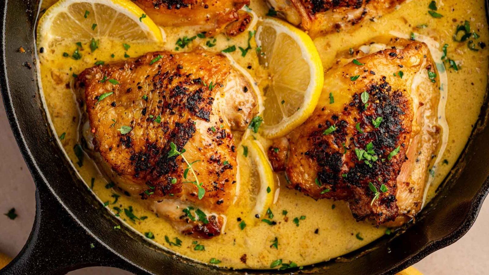 Four browned chicken thighs in a creamy lemon sauce, garnished with parsley and lemon slices, cooking in a cast iron skillet.