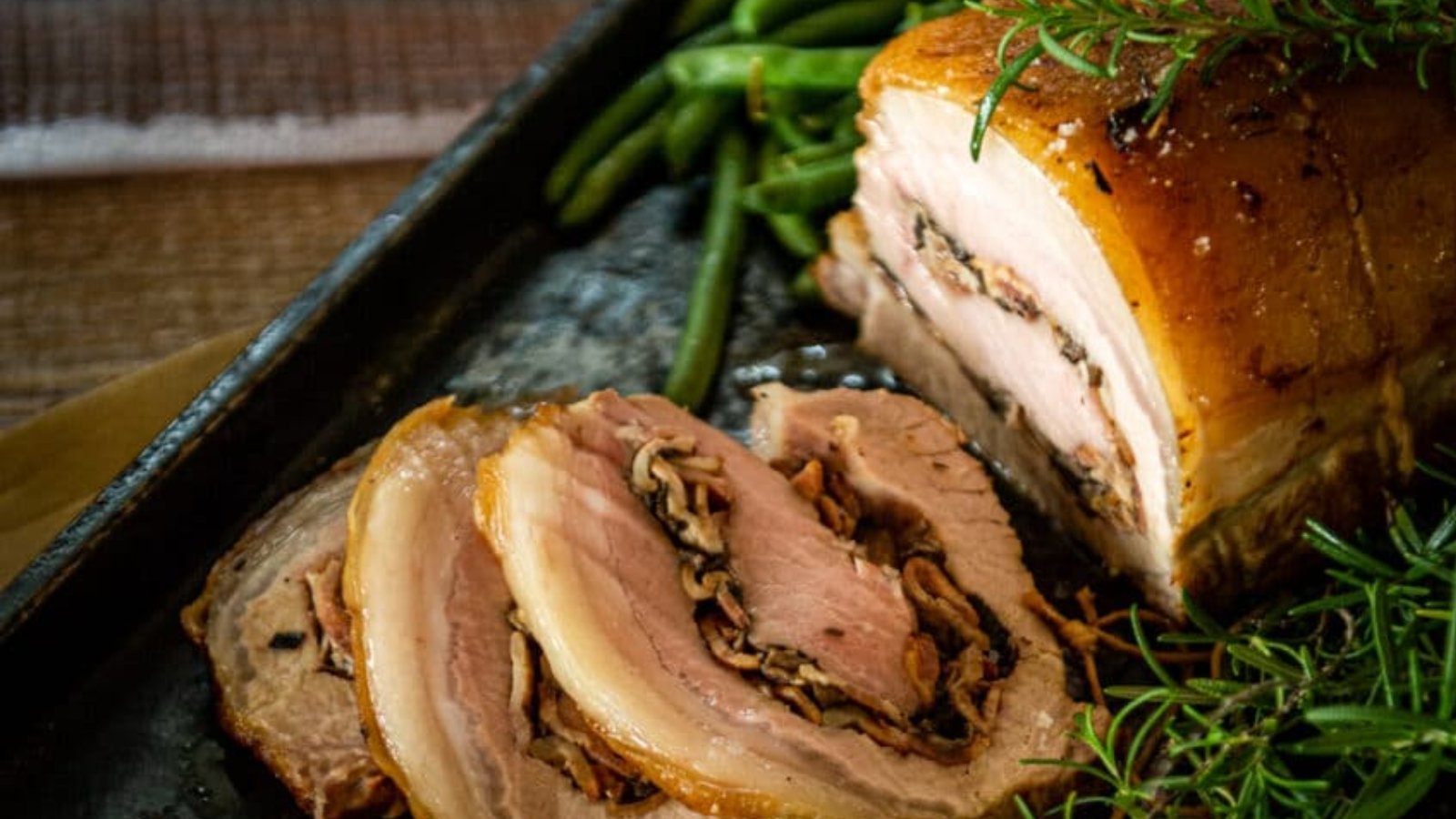 A close-up of sliced rolled pork roast with a herb and mushroom filling, served with green beans and garnished with fresh rosemary.