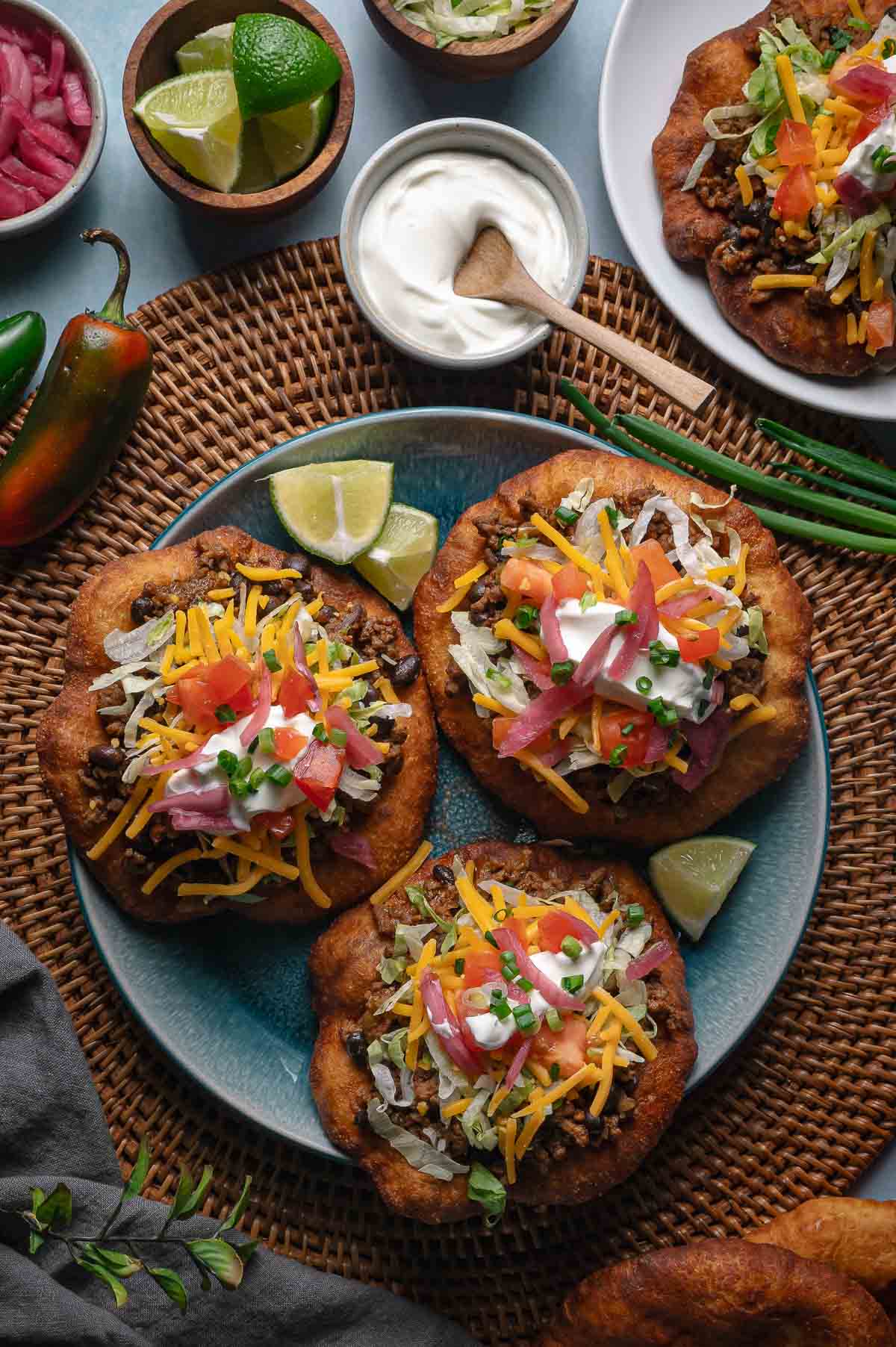 Three fry bread tacos topped with ground meat, shredded cheese, lettuce, tomatoes, pickled onions, and sour cream on a blue plate, surrounded by lime wedges and condiments.