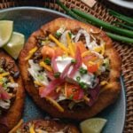 A plate of fry bread topped with ground taco meat, shredded lettuce, cheddar cheese, diced tomatoes, pickled onions, sour cream, and chopped chives, with lime wedges on the side.