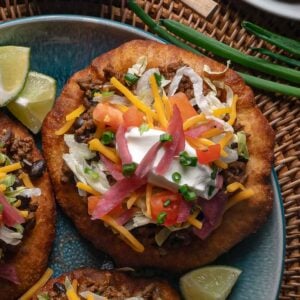 A plate of fry bread topped with ground taco meat, shredded lettuce, cheddar cheese, diced tomatoes, pickled onions, sour cream, and chopped chives, with lime wedges on the side.