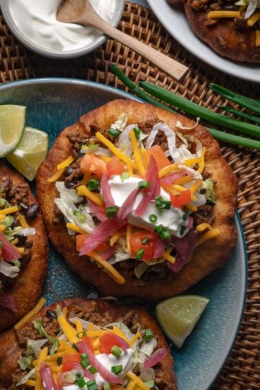 A plate of fry bread topped with ground taco meat, shredded lettuce, cheddar cheese, diced tomatoes, pickled onions, sour cream, and chopped chives, with lime wedges on the side.