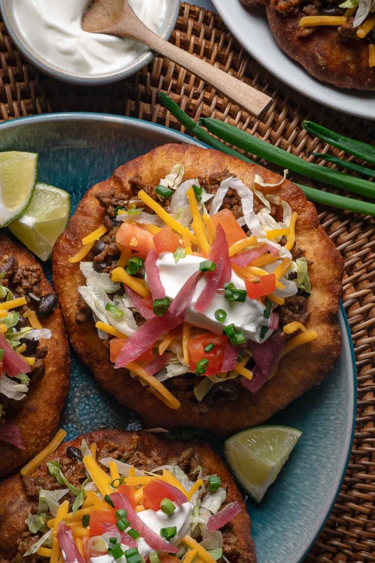 A plate of fry bread topped with ground taco meat, shredded lettuce, cheddar cheese, diced tomatoes, pickled onions, sour cream, and chopped chives, with lime wedges on the side.