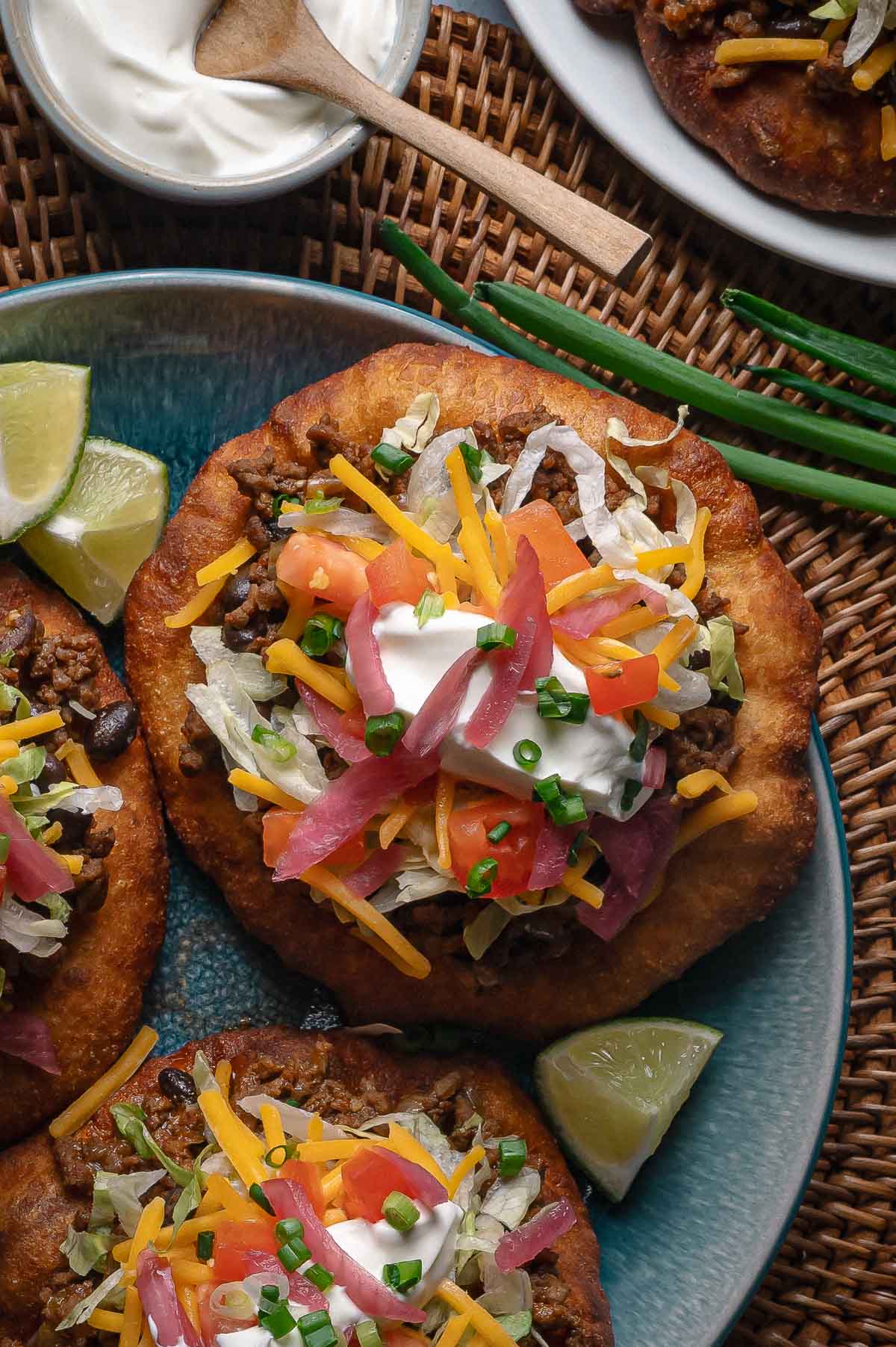 A plate of fry bread topped with ground taco meat, shredded lettuce, cheddar cheese, diced tomatoes, pickled onions, sour cream, and chopped chives, with lime wedges on the side.