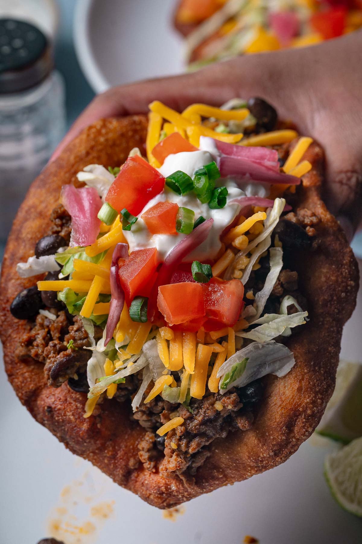 A hand holds a fluffy Navajo taco filled with ground beef, beans, shredded cheese, lettuce, diced tomatoes, sour cream, and green onions.