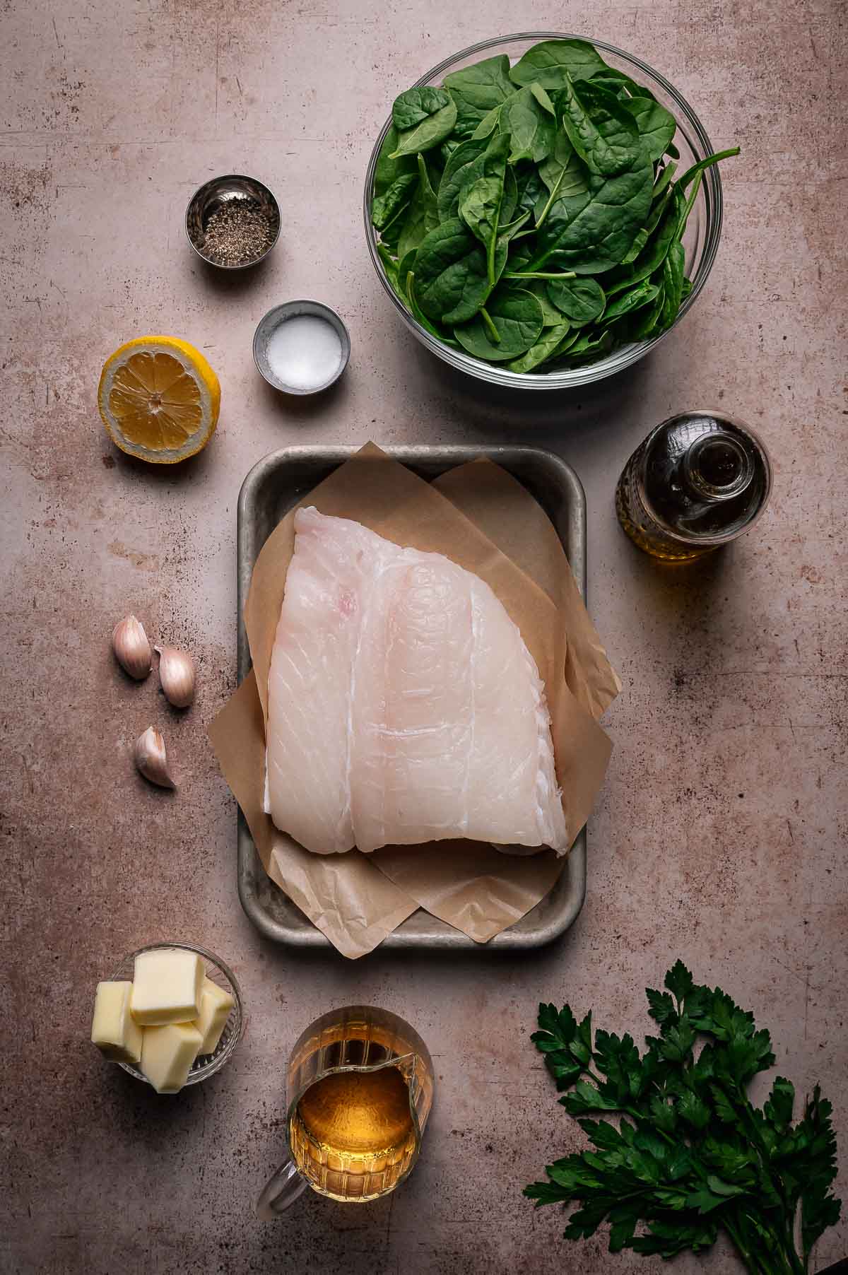 Raw halibut fillets on parchment paper with spinach, butter, garlic, lemon, parsley, oil, salt, pepper, and a small glass of white wine arranged on a counter.