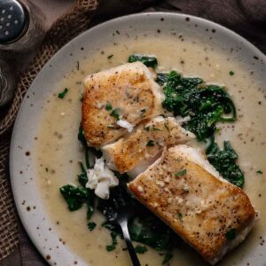 A plate of seared halibut fillets served over sautéed spinach with a creamy sauce, garnished with chopped herbs. A fork rests on the plate.