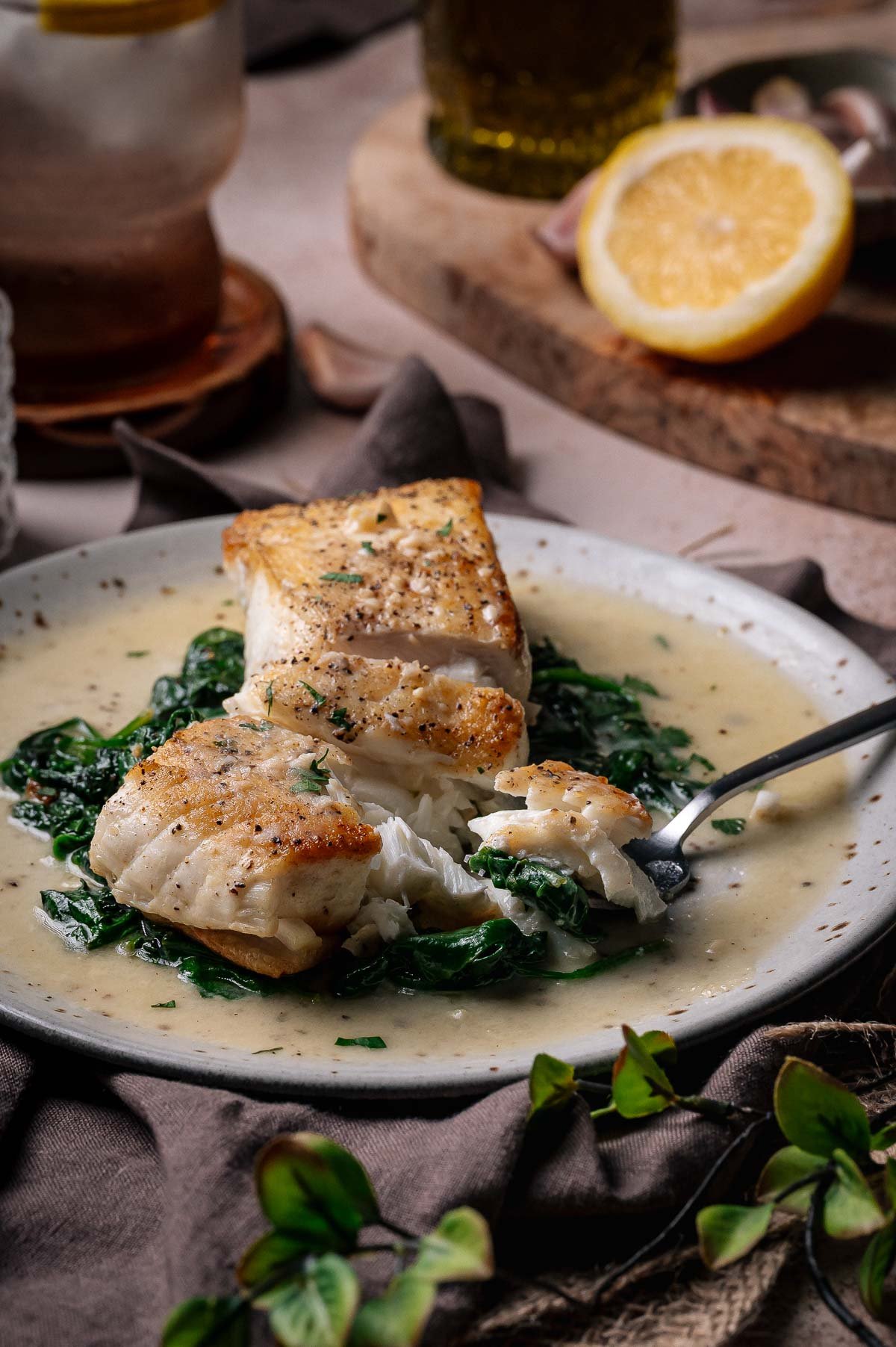 A plate with seared halibut fillets on a bed of sautéed spinach, topped with creamy sauce, with a fork taking a bite. A lemon half and other ingredients are in the background.