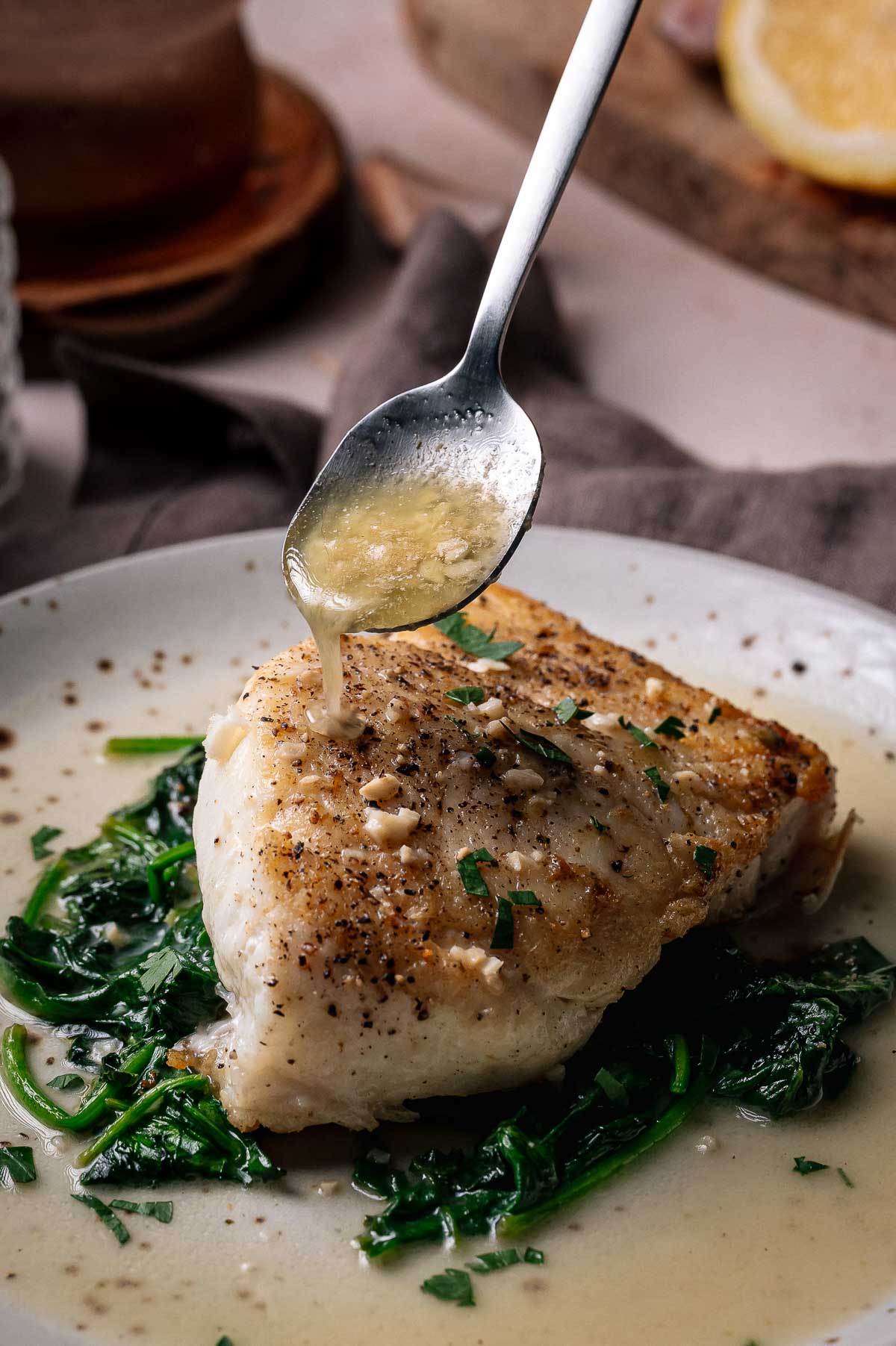 A piece of seasoned halibut sits on sautéed spinach as a spoon drizzles sauce over it on a white plate.