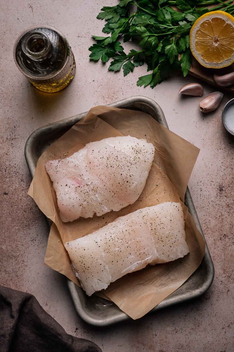 Two raw halibut fillets on parchment paper in a metal tray, seasoned with pepper, with olive oil, parsley, lemon, and garlic beside them.
