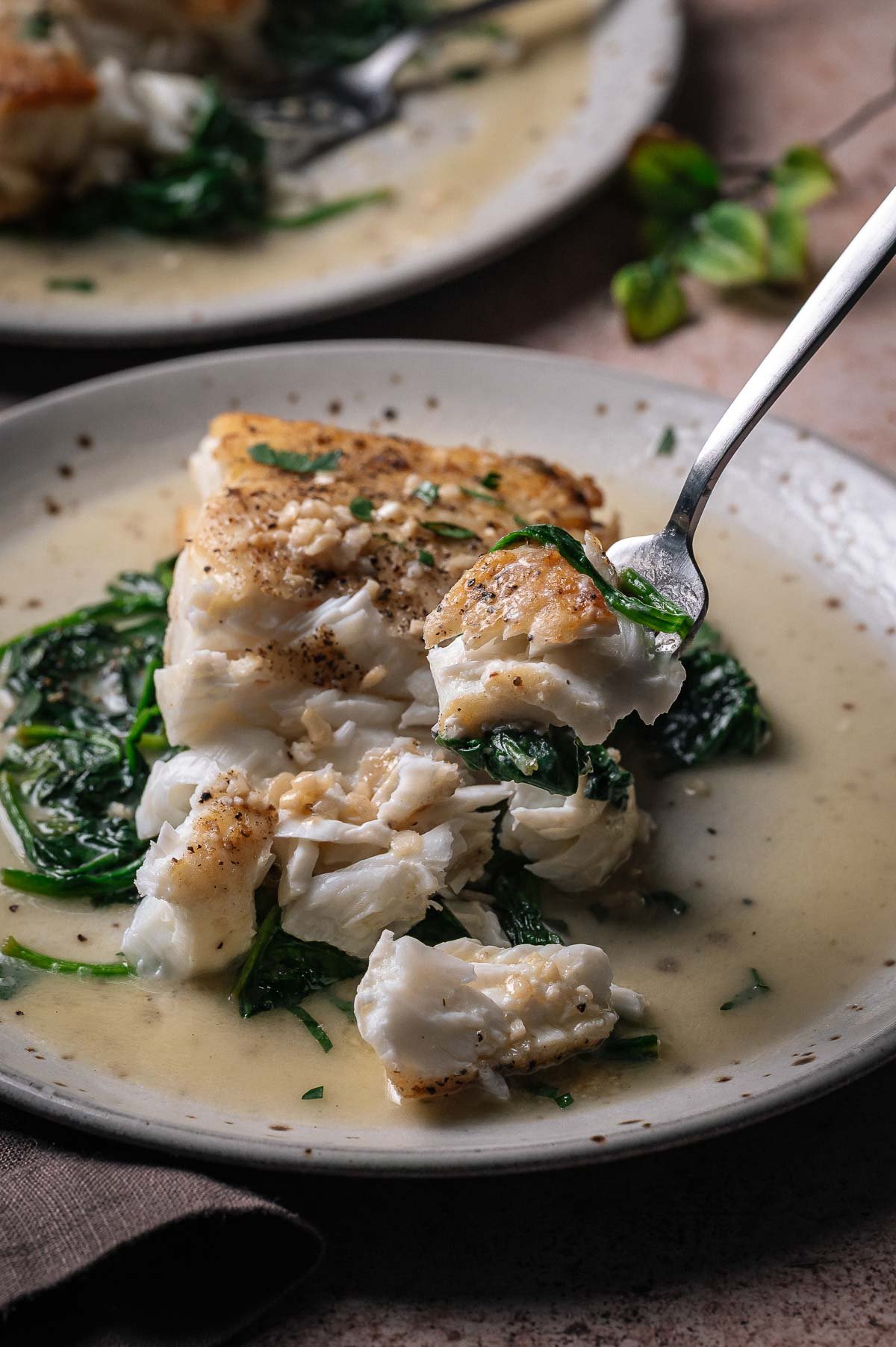 A fork lifts a piece of cooked white fish with wilted spinach in a creamy sauce from a plate.