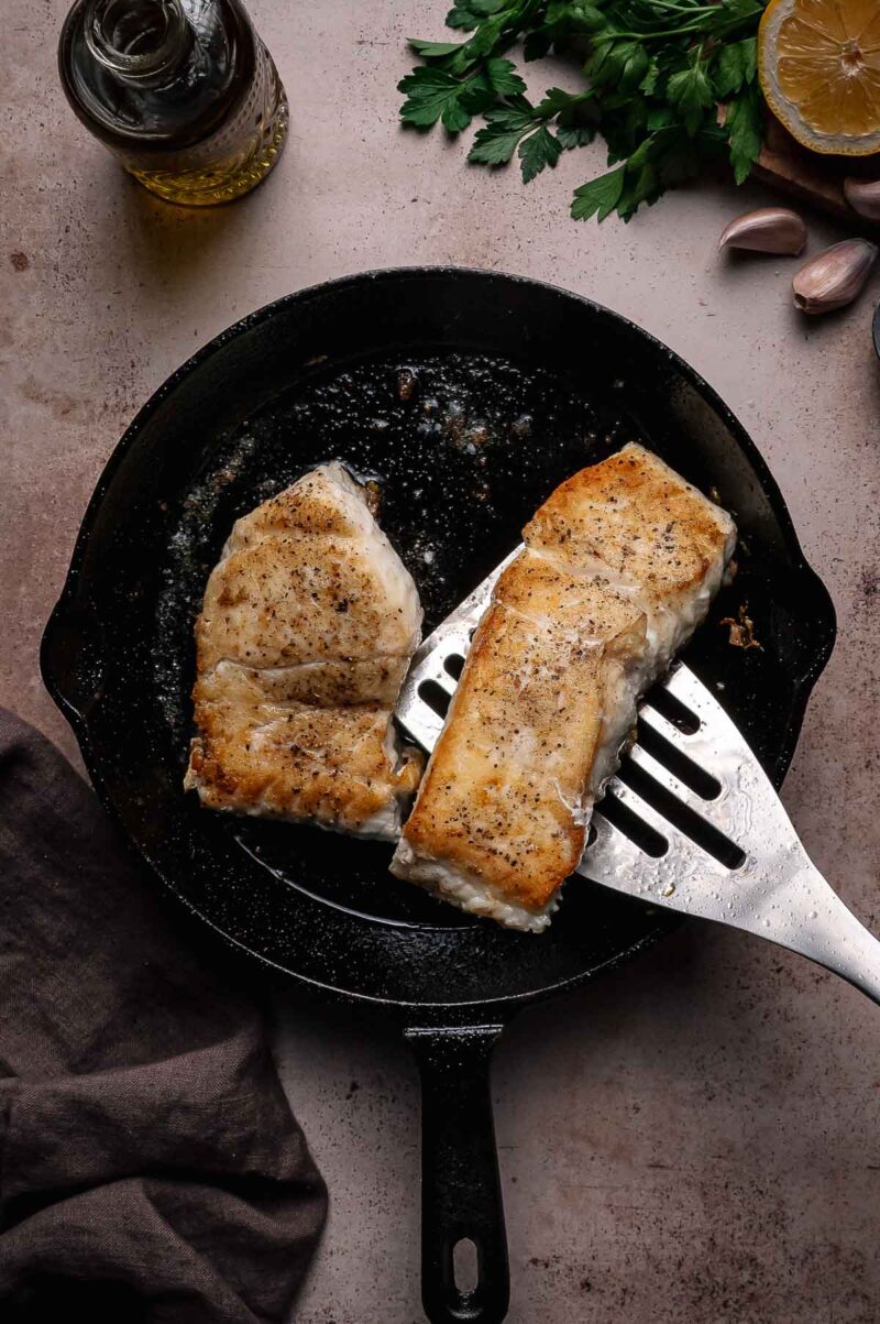 Two seared halibut fillets in a cast iron skillet, one being lifted by a metal spatula; parsley, lemon, garlic, and olive oil are nearby.
