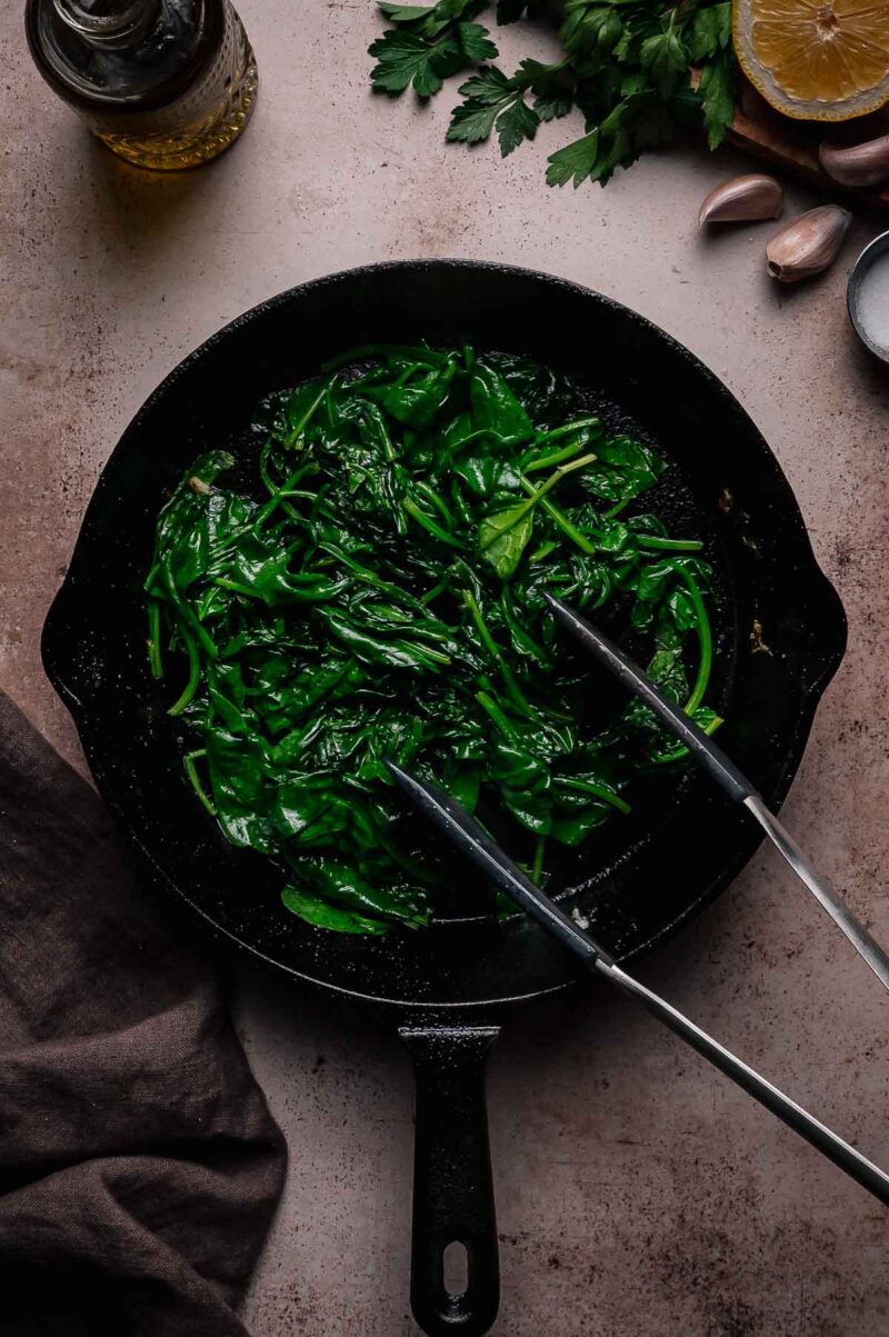 A cast iron skillet with sautéed spinach and metal tongs, surrounded by garlic, lemon, fresh herbs, olive oil, and a brown napkin.