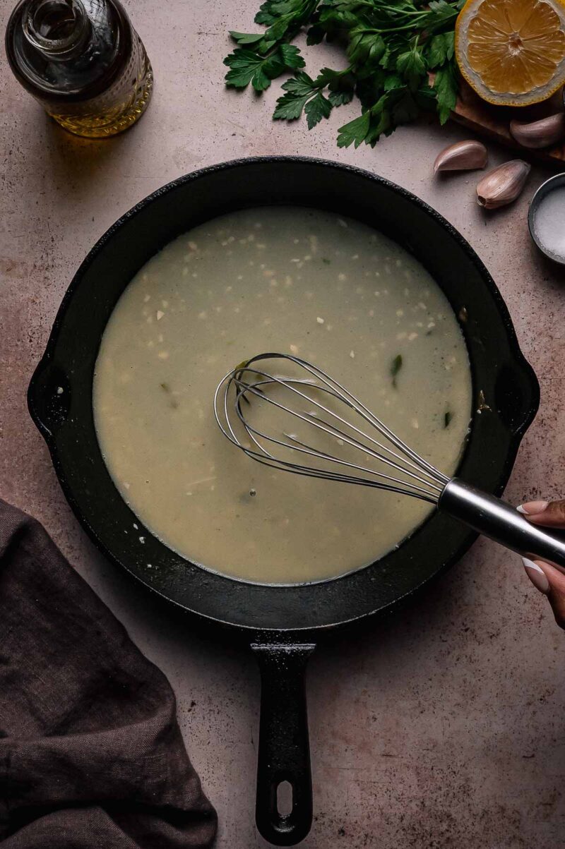 A hand whisking a light-colored sauce in a black skillet, surrounded by parsley, garlic, a lemon half, salt, and a bottle of oil on the table.