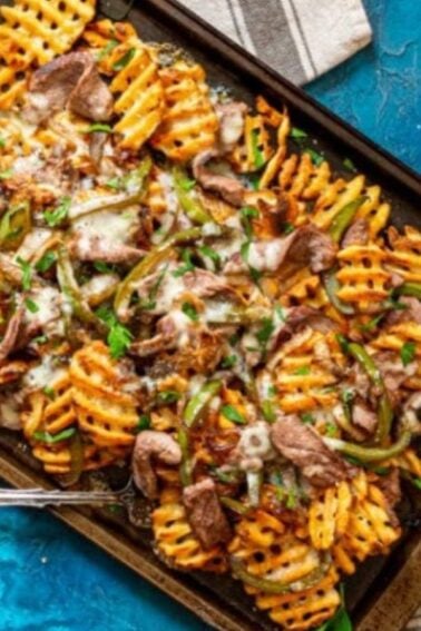 A baking tray with waffle-cut fries, sliced beef, green peppers, melted cheese, and herbs on a blue surface, with a spoon and napkins beside it.