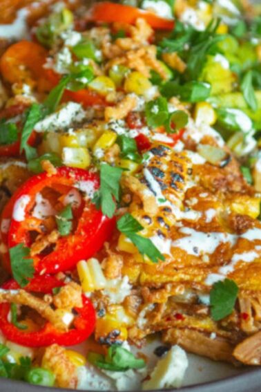A plate of enchiladas topped with sliced red peppers, fresh herbs, white sauce, and lime wedges, with a fork on the side.