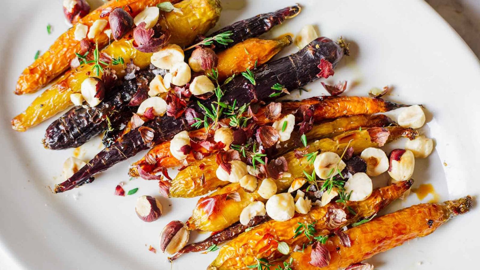 A plate of roasted carrots in various colors, topped with chopped hazelnuts and sprigs of fresh thyme. The dish is presented on a white plate.