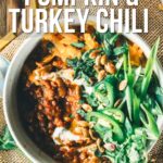 Overhead view of a bowl of roasted pumpkin and turkey chili topped with greens, cheese, and pumpkin seeds, placed on a textured surface with herbs and tortilla chips nearby.