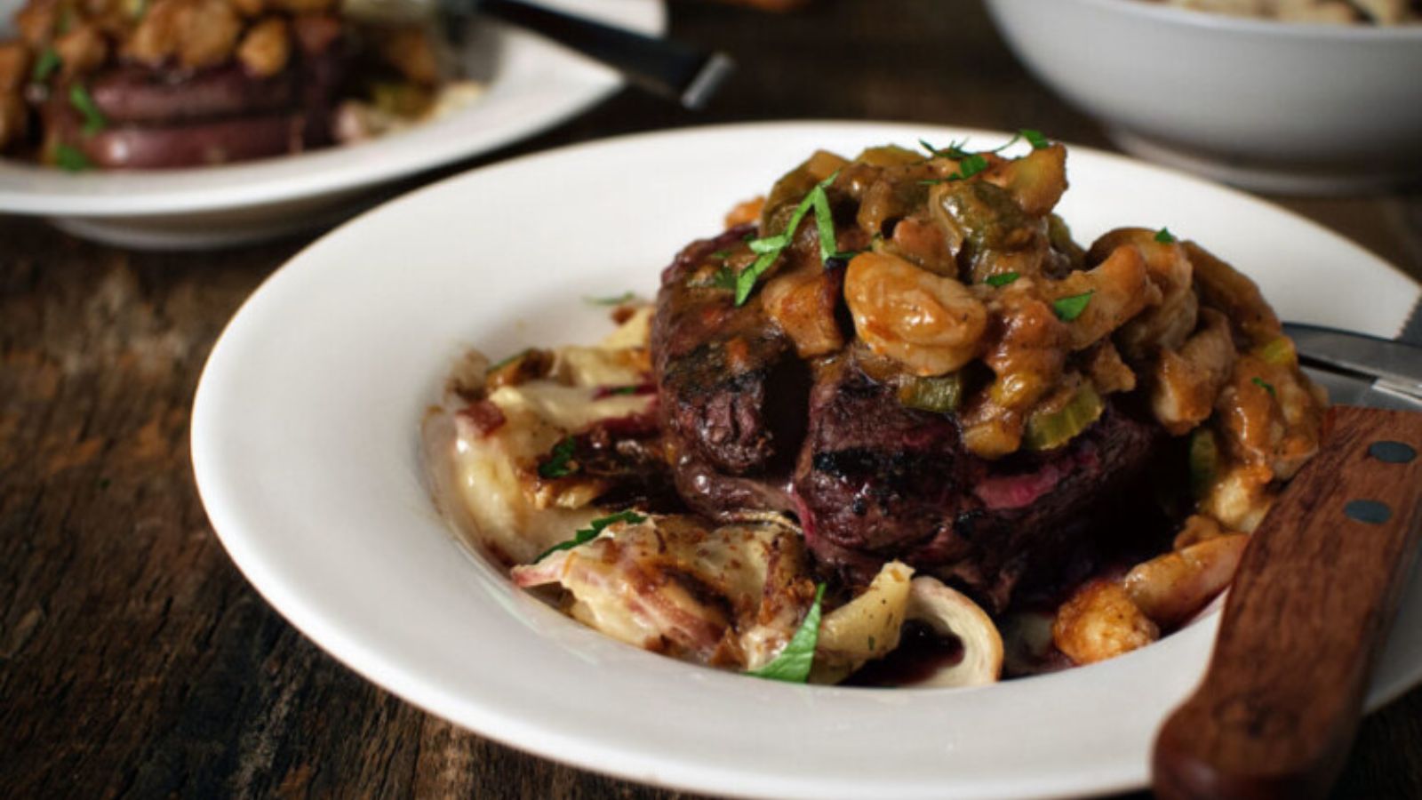 A plate with a cooked steak topped with a chunky sauce, served over creamy pasta, garnished with chopped herbs. A knife is placed on the plate edge.