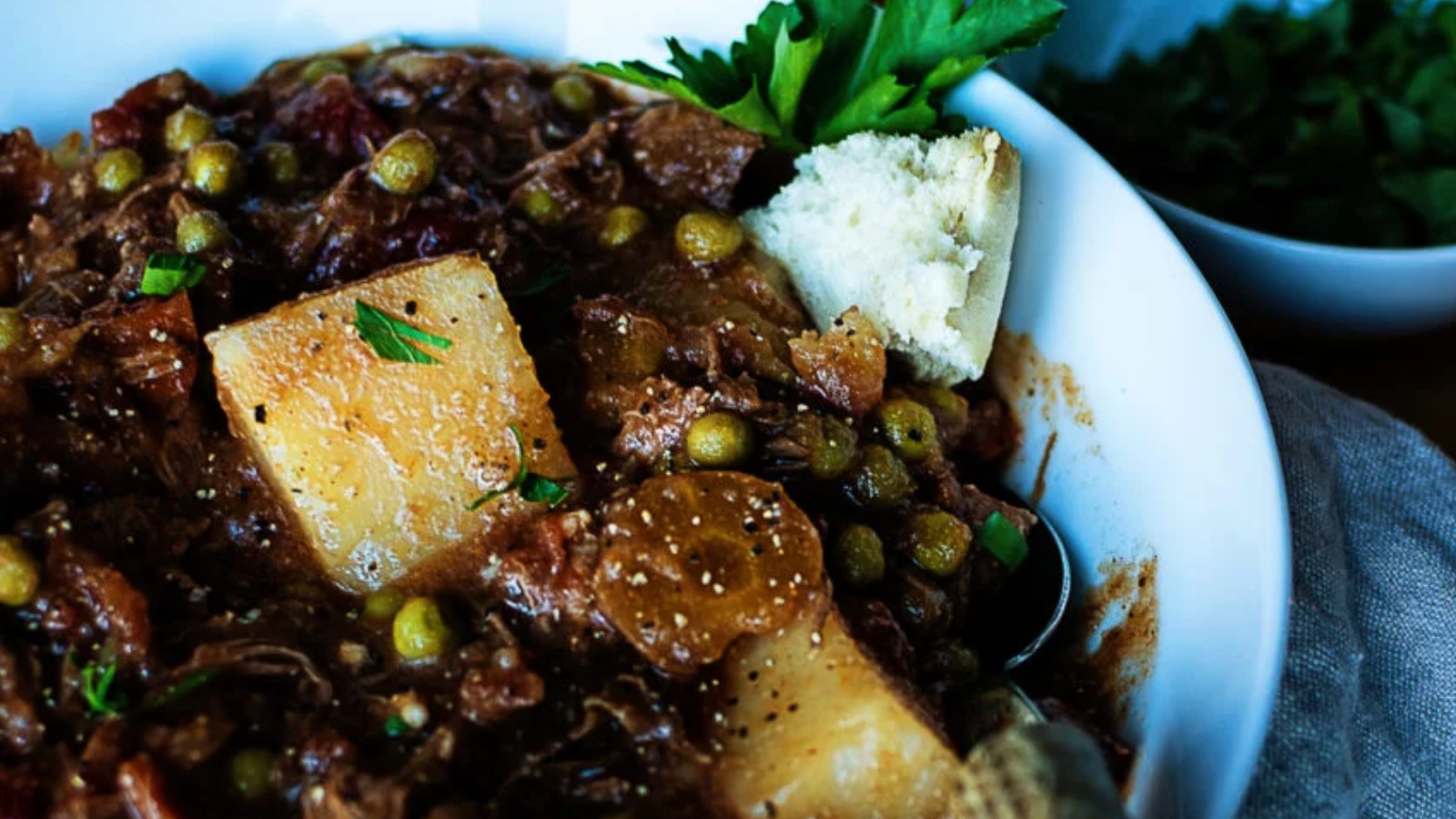 A close-up of a bowl of beef stew with large potato chunks, peas, herbs, and a piece of bread on the side.