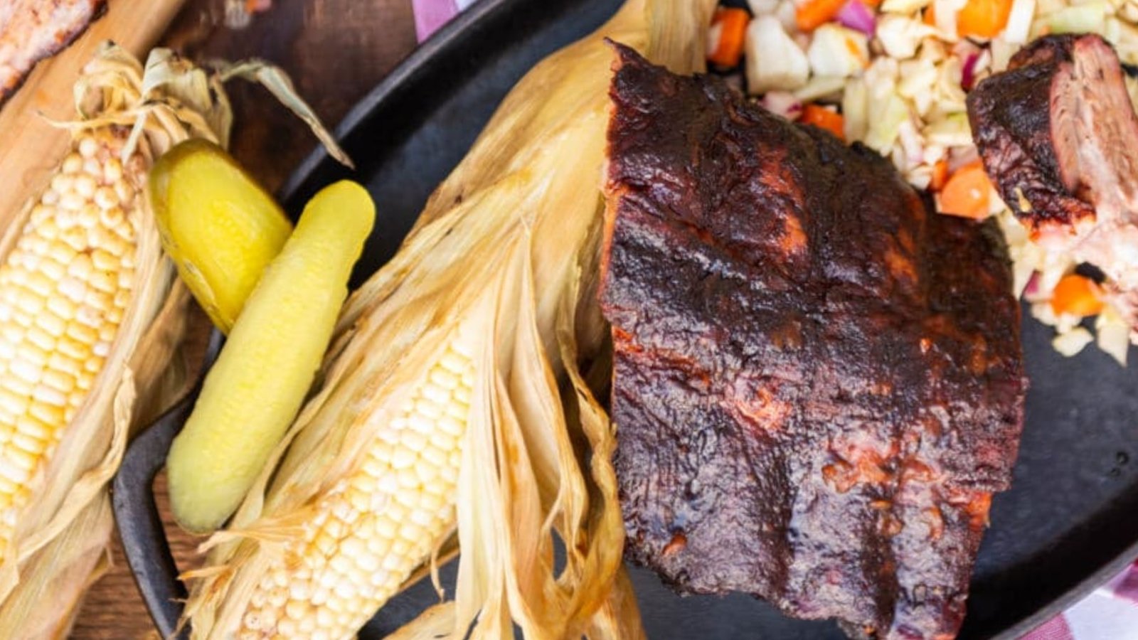 A plate with grilled corn on the cob, pickles, coleslaw, and a portion of barbecued ribs.
