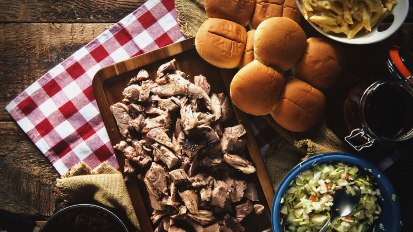 Overhead view of pulled pork on a wooden board, hamburger buns, coleslaw, pasta, and a red-and-white checkered cloth on a wooden table.