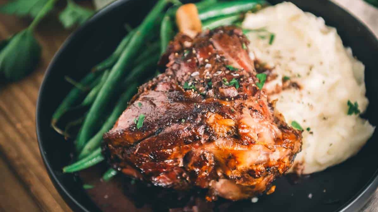 A plate with braised lamb shank, mashed potatoes, and green beans, garnished with herbs.