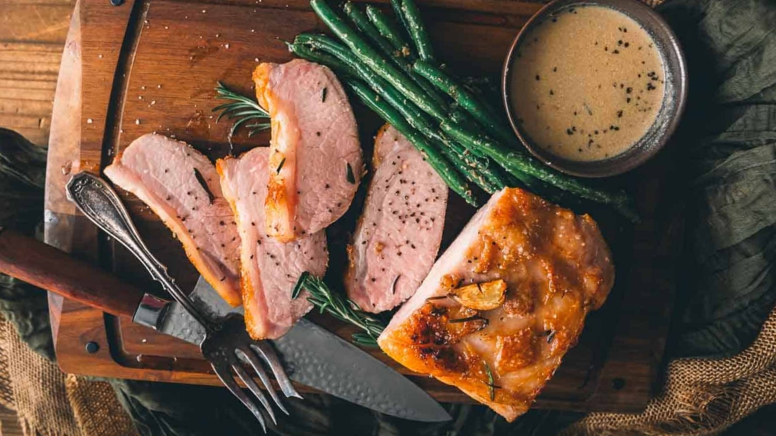 Sliced roasted pork with green beans, a fork and knife, and a bowl of creamy sauce on a wooden cutting board.
