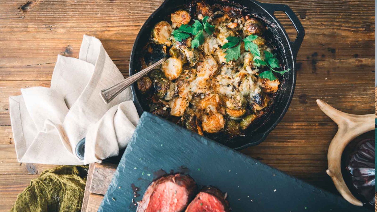 A cast iron skillet with roasted potatoes and herbs beside a slate board with sliced cooked meat on a wooden table.