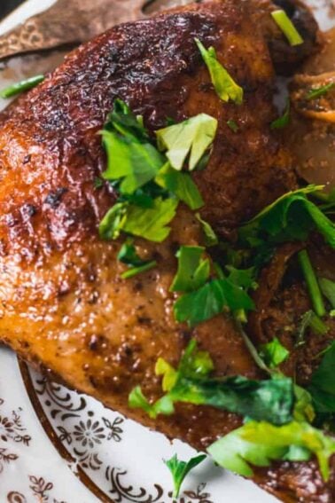 A cooked chicken leg with browned skin served on a patterned plate alongside sliced potatoes, garnished with fresh chopped parsley.