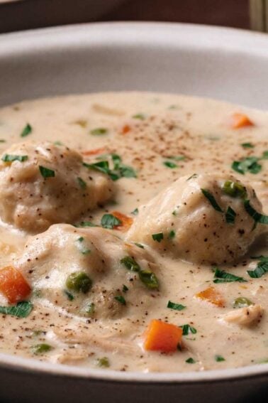 A bowl of creamy chicken and dumplings soup with carrots, peas, and herbs, served with a spoon.