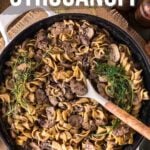 A skillet filled with ground beef stroganoff made with noodles, mushrooms, and herbs, with a wooden spoon resting in the dish. Text overlay reads "How to Make Ground Beef Stroganoff.