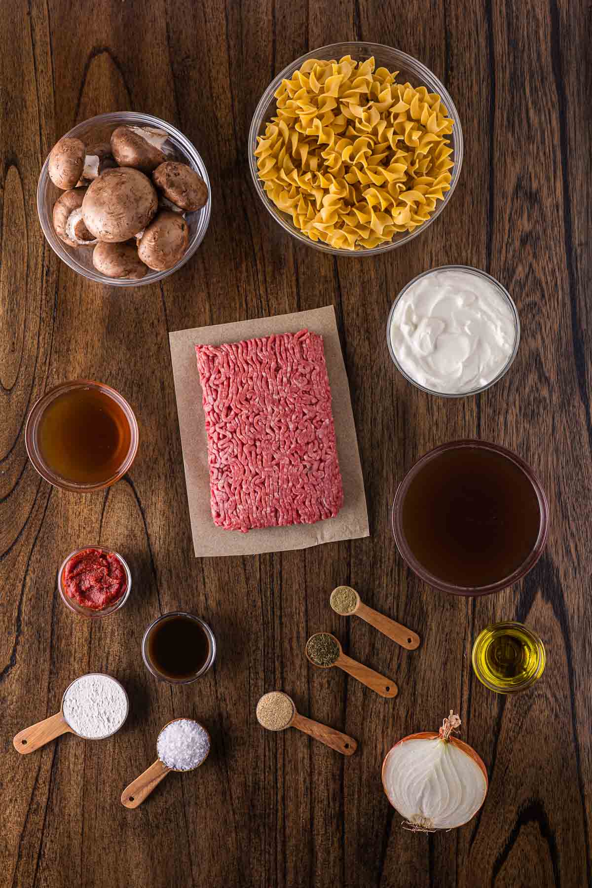 Ingredients for a beef stroganoff recipe arranged on a wooden table, including ground beef, egg noodles, mushrooms, sour cream, broth, spices, flour, tomato paste, onion, and oil.