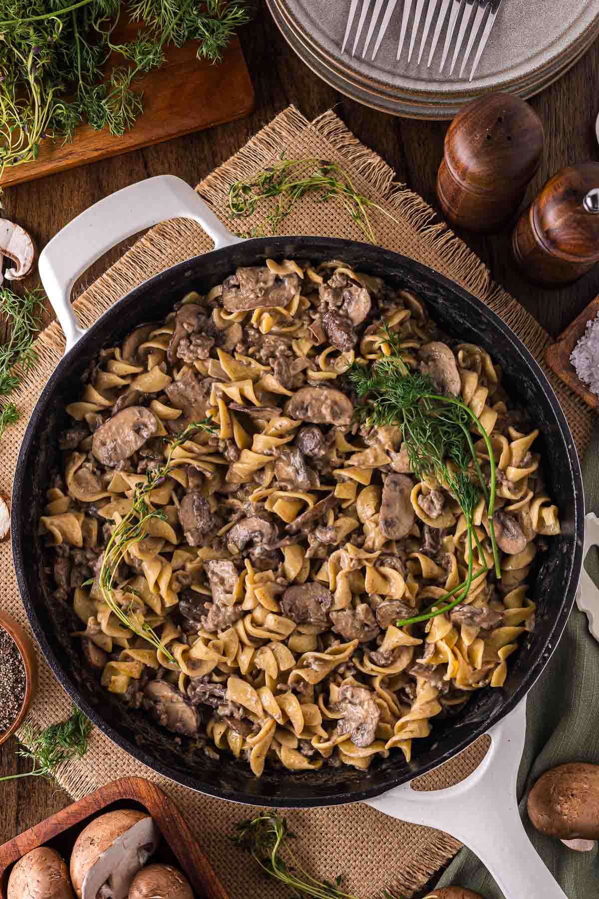 A skillet filled with beef stroganoff made with egg noodles, mushrooms, ground beef, and garnished with fresh dill; various ingredients and utensils surround the pan.