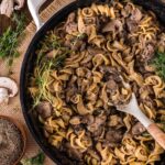 A skillet filled with ground beef stroganoff and egg noodles, garnished with fresh herbs, with a wooden spoon resting inside. Surrounding the skillet are mushrooms, herbs, and pepper.
