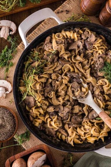 A skillet filled with ground beef stroganoff and egg noodles, garnished with fresh herbs, with a wooden spoon resting inside. Surrounding the skillet are mushrooms, herbs, and pepper.