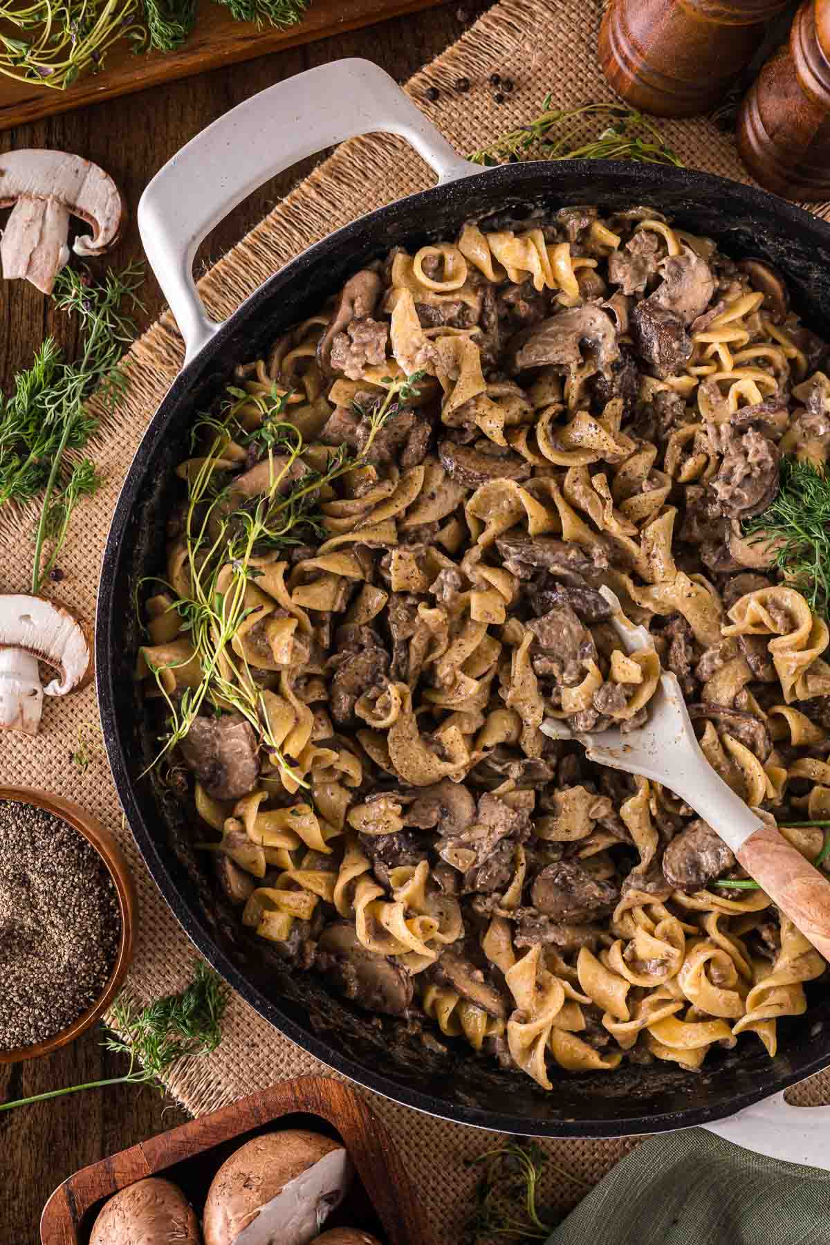 A skillet filled with ground beef stroganoff and egg noodles, garnished with fresh herbs, with a wooden spoon resting inside. Surrounding the skillet are mushrooms, herbs, and pepper.